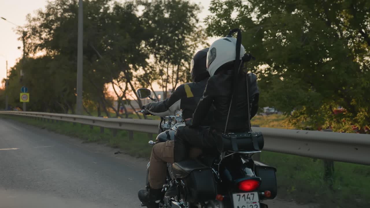 Motorcycle rider with passenger wearing helmets and leather jackets travels along asphalt road during sunset, surrounded by cars, guardrails, and roadside trees