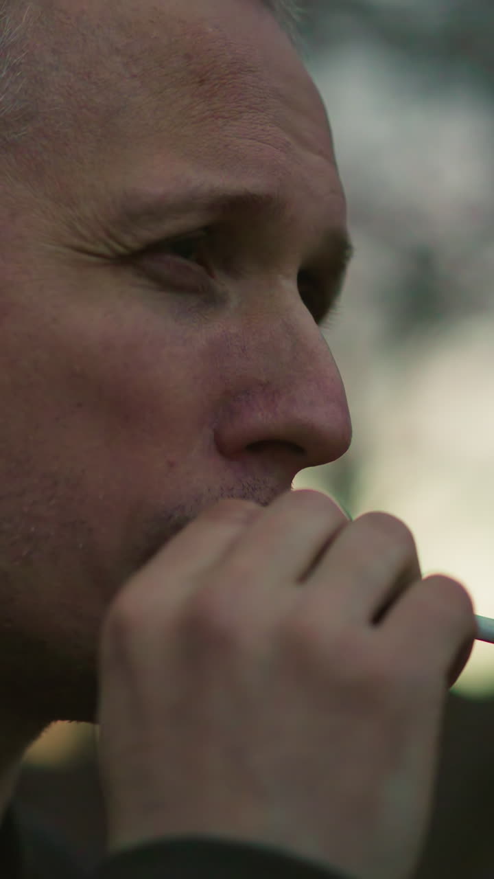 близкий боковой вид человека в зеленой куртке, выдыхающего дым через нос и рот в сумерках. размытый фон с деревьями и мягким светом