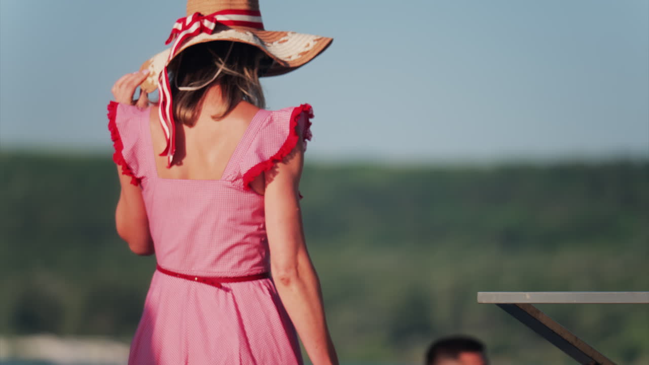 Woman in a red gingham dress and wide hat with a ribbon walking on a terrace
