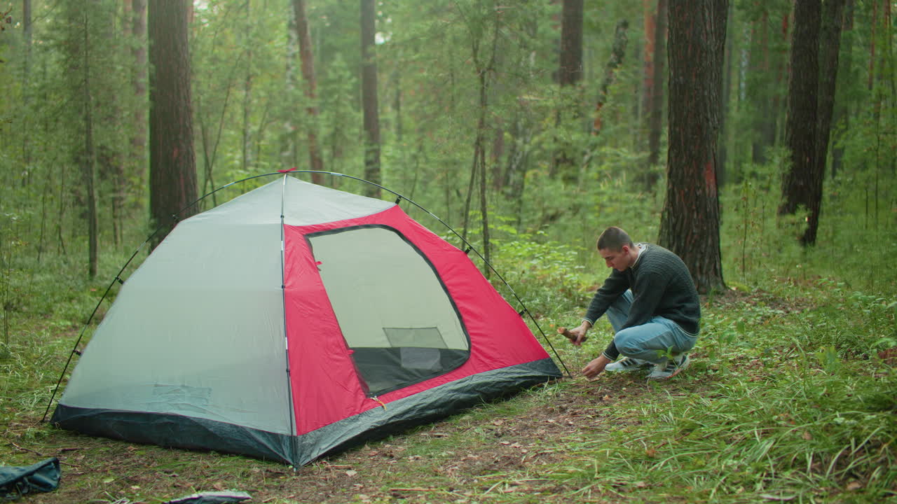 man crouches beside tent in green forest holding wood stick and driving peg into ground with focused expression, surrounded by trees and natural light during outdoor camping activity