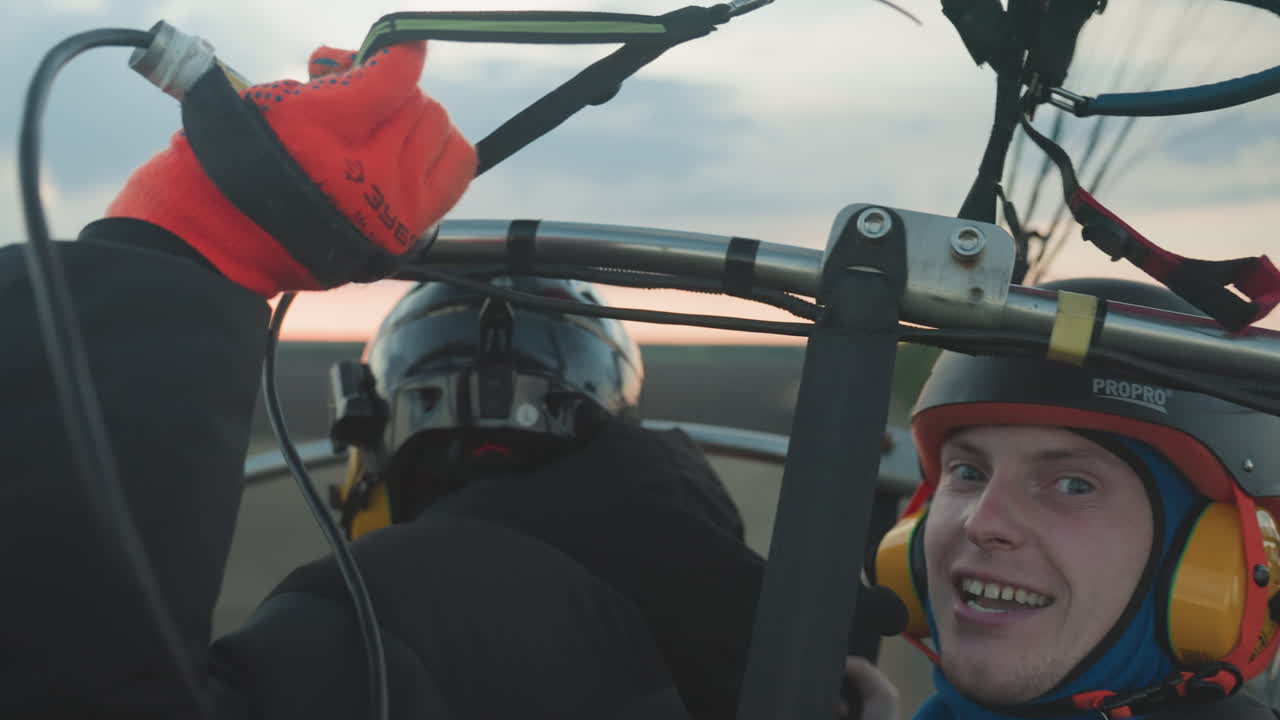 closeup of excited pilot in checkered jacket helmet and headset gripping paraglider frame with wide eyes and open mouth against blurred farmland and pastel sunset sky before launching