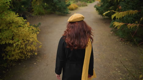 back view of a lady with a yellow beret and muffler, adjusting her hair as she strolls through a vibrant garden filled with autumn colors