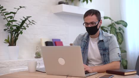 joven caucásico se queda en casa trabajando desde casa usando una máscara protectora usando computadora portátil e internet. oficina en casa acogedora, lugar de trabajo pandemia de coronavirus, cuarentena de covid-19. trabajo remoto, freelance