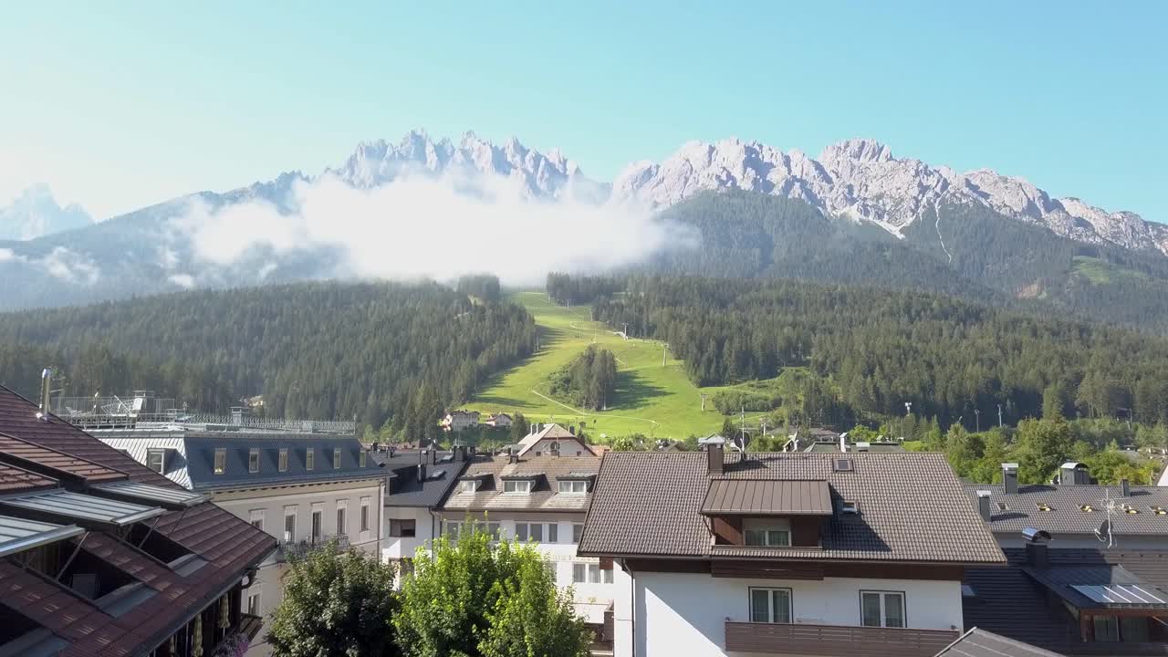 volar sobre un pueblo de estación de esquí en verano con una pista de esquí verde en el fondo, innichen, tirol del sur, italia