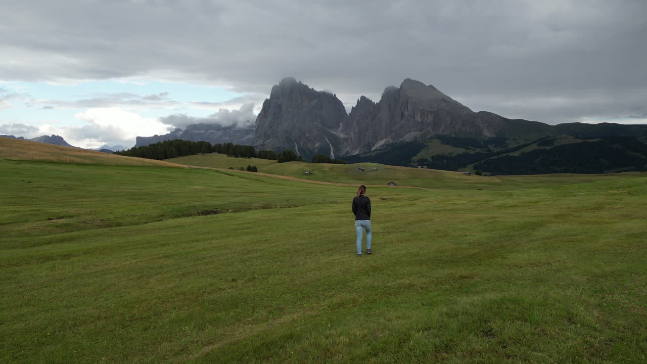 Female hiker walking through verdant meadow, approaching iconic Sciliar massif in Alpe di Siusi, showcasing stunning Alpine landscape and serene natural beauty of South Tyrol, Italy