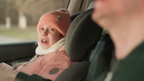 a young girl, dressed warmly in a pink beanie and jacket, is sitting in the front passenger seat of a car, singing with enthusiasm. beside her, a man, slightly blurred in the background