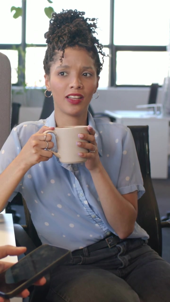 Vídeo vertical: Mulher sentada, segurando uma caneca bege, reage enquanto colega de trabalho mostra smartphone no escritório.