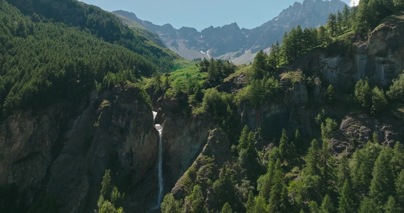Il drone si avvicina alla spettacolare Cascata di Barliard che scende a cascata da una scogliera, incorniciata da una pineta e imponenti cime di granito sotto un cielo estivo limpido e azzurro in Italia.