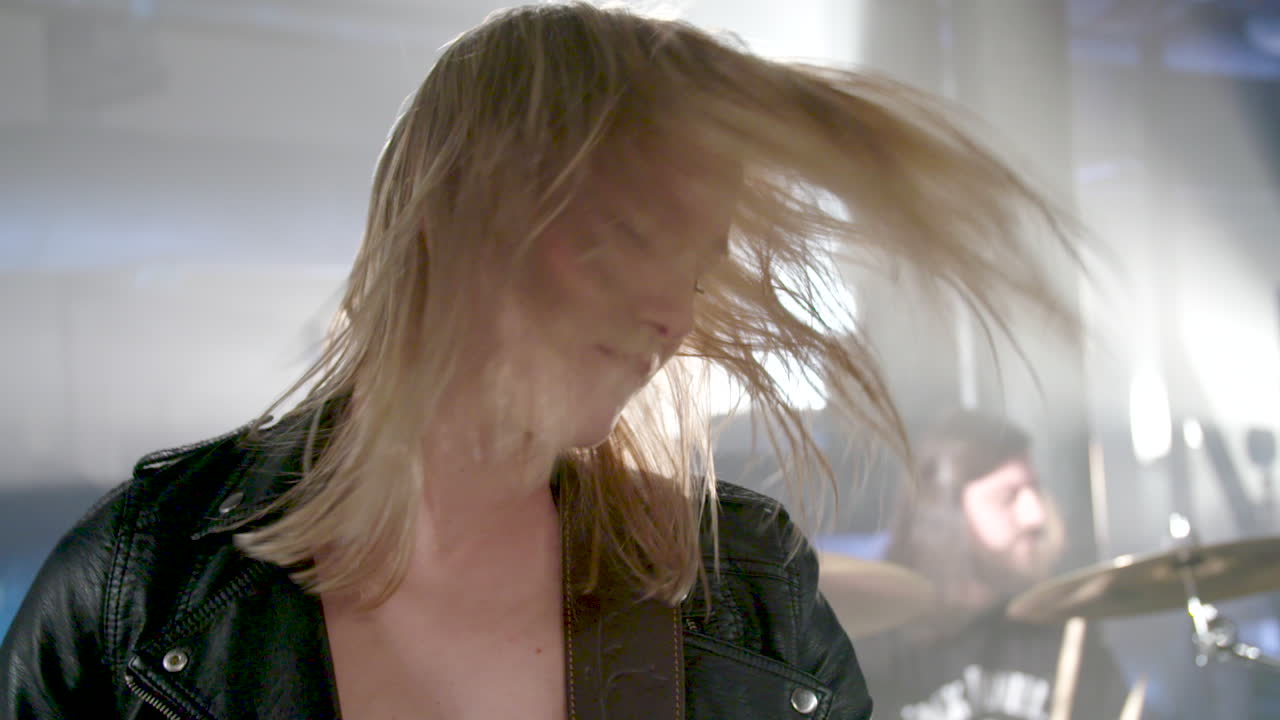 Red lipped girl playing bass swings her head on stage to the music (close-up, slowmotion). She wears a leather jacket. A drummer performs in the background.