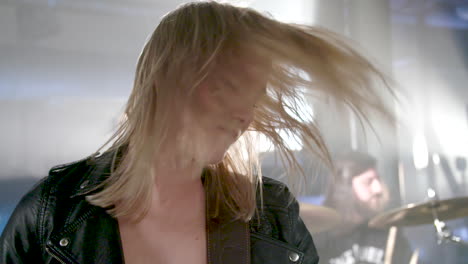 red lipped girl playing bass swings her head on stage to the music (close-up, slowmotion). she wears a leather jacket. a drummer performs in the background.