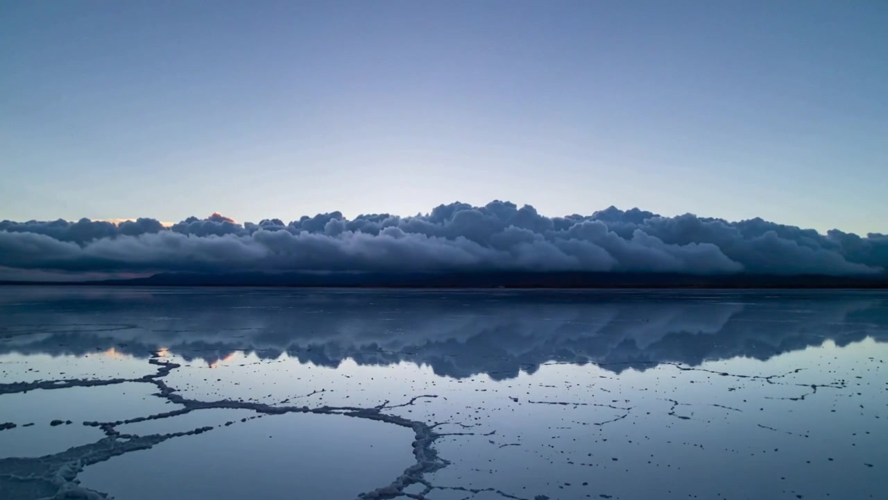 Skiftende lang lav skybanke og refleksion ændrer sig med solopgangsglød, der viser sig på en saltslette.