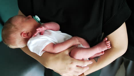 little caucasian baby in a white bodysuit in mom's hands. lovely child with a pacifier sleeping. close up.