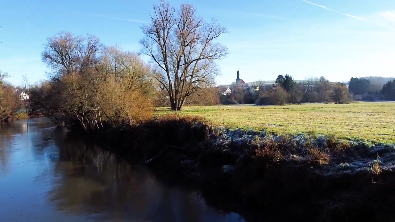 Et filmisk udsyn over en stille flod, der snor sig gennem frostklare marker med en kirke og hyggelige huse i det fjerne. En fredfyldt vintermorgen med en klar blå himmel