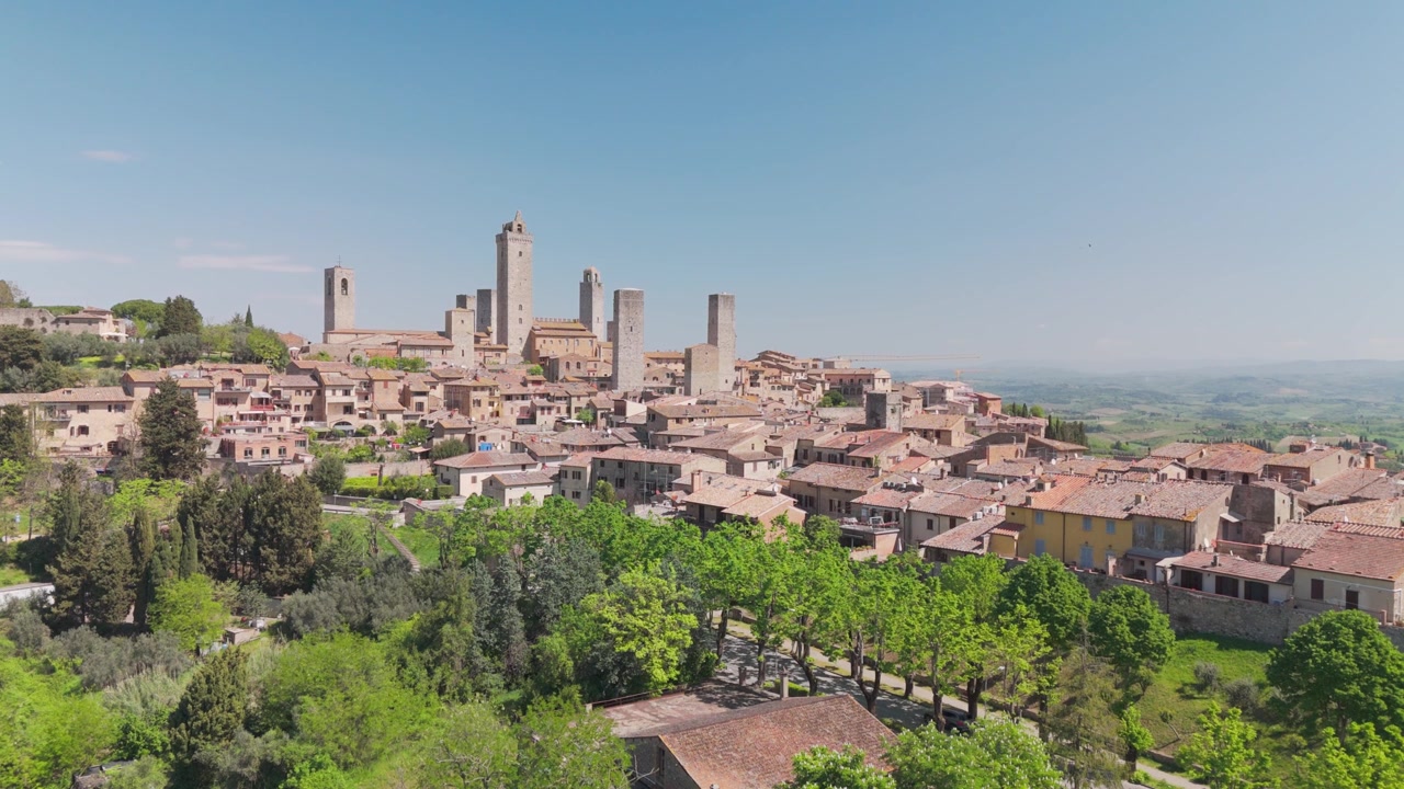 Drone stazionario con una vista diretta di San Gimignano, Toscana, Italia, con una fila di alberi in primo piano e un'auto che la attraversa, rivelando la città collinare medievale sotto una calda luce toscana