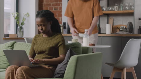 girl working on laptop computer sitting on sofa while her male roommate cleaning up after breakfast