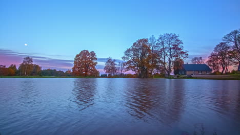 Der-Mond-Geht-Unter,-Während-Der-Sonnenaufgang-Die-Herbstfarbenen-Bäume-An-Einem-See-Und-Einer-Hütte-Beleuchtet---Zeitraffer