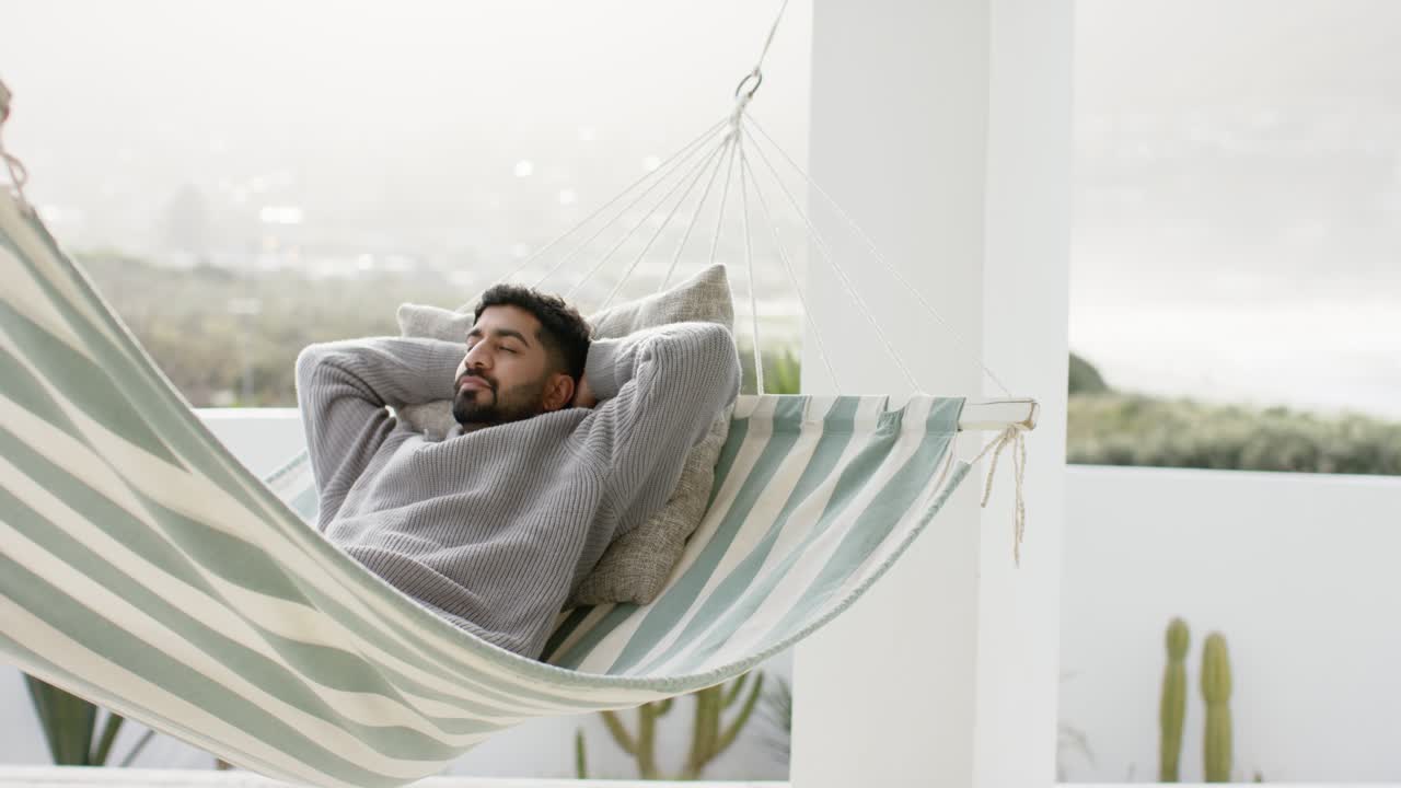 Hombre indio descansando en una hamaca en un balcón, apoyado en un cojín y respirando con calma para relajarse