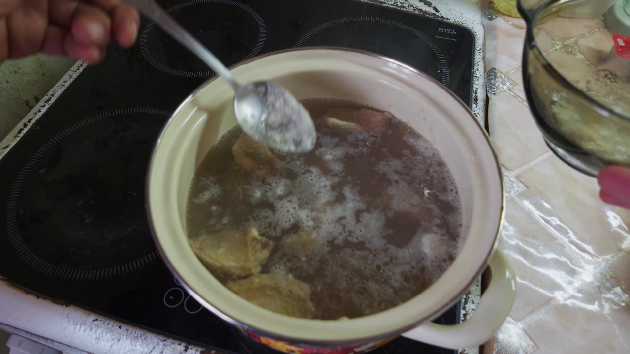 Una mano usa un cucchiaio per togliere la schiuma da una pentola di brodo di carne che sobbolle su un fornello elettrico. Il liquido in leggera ebollizione viene depurato dalle impurità mentre un coperchio di vetro è appoggiato lì vicino sul piano di lavoro.