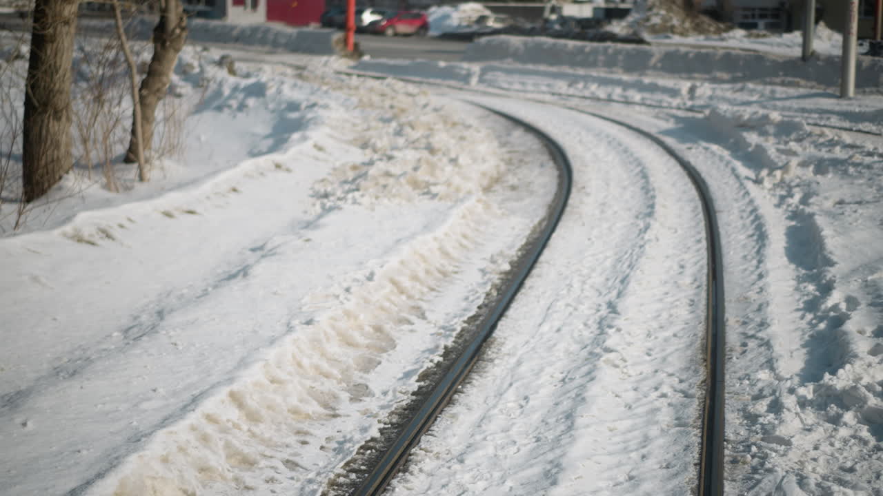 window view from moving train shows curved tracks cut through winter snow, trees lining route, frosty embankment, smooth motion conveying cold commute along city edge toward distant junction