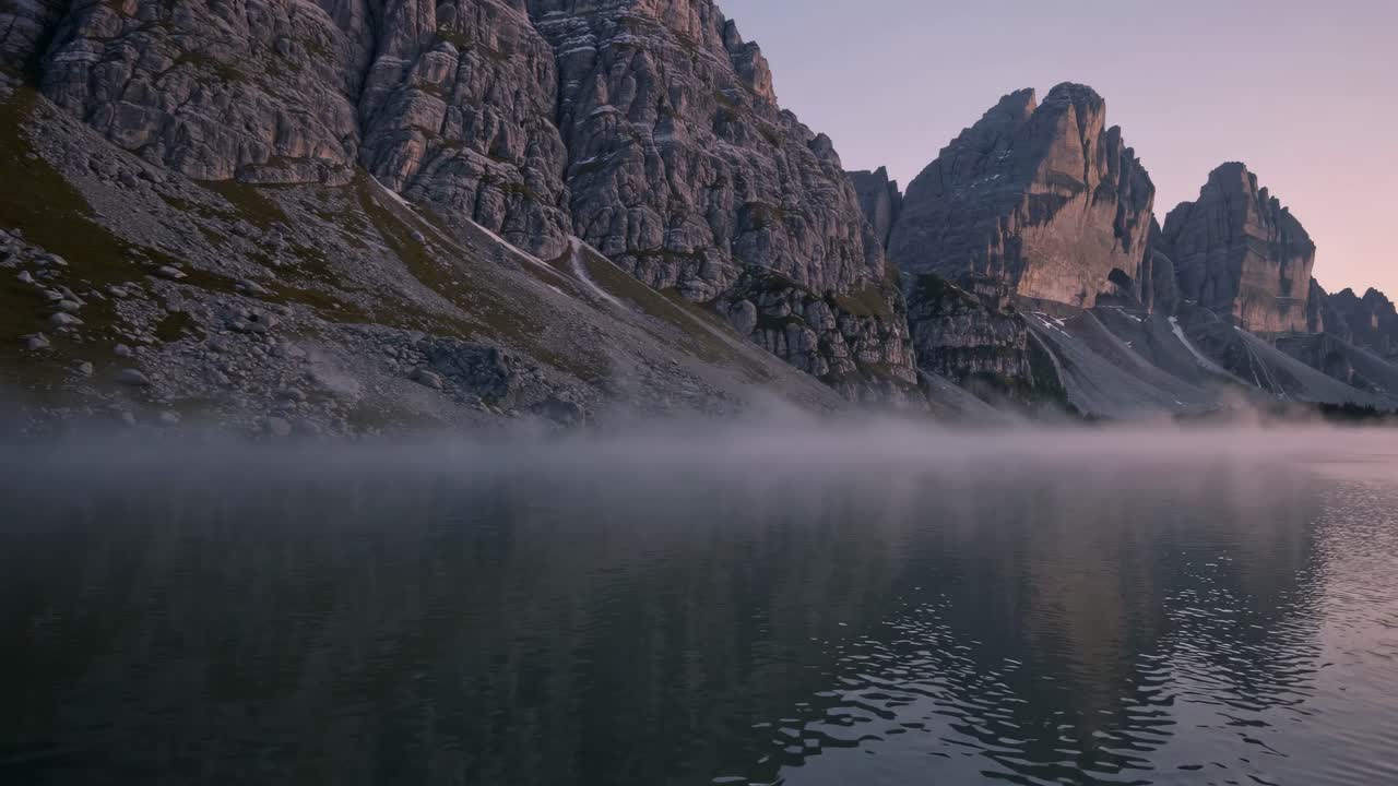 A serene video captures a low-angle view of misty mountains reflecting in a calm lake at dawn