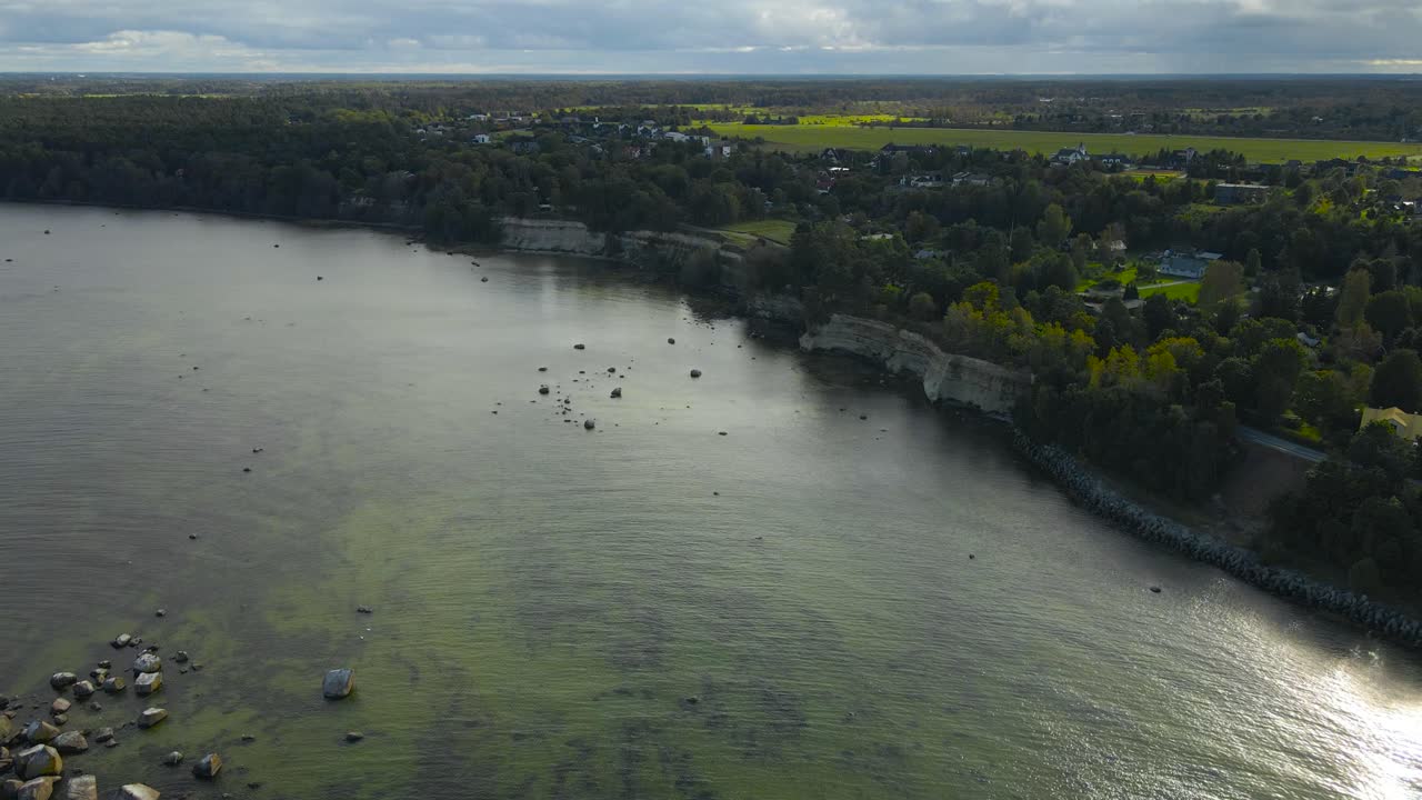 Aerial drone footage flying above a gorgeous limestone cliff bank during summer sunny day in Tilgu Estonia, wtih treetops and small private houses visible. Horizon seen in the background, farm lands