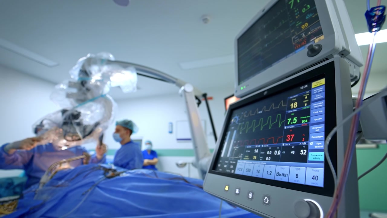 I monitor del ventilatore polmonare mostrano i segni vitali del paziente. Il medico al tavolo operatorio sposta il microscopio coperto di plastica. Sfondo sfocato.