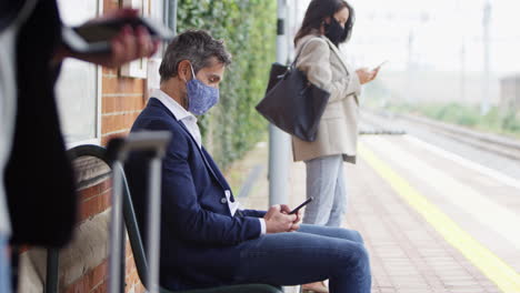 business commuters on railway platform with mobile phones wearing ppe face masks during pandemic