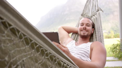 a young caucasian man relaxes in a hammock with a tablet in the backyard at home