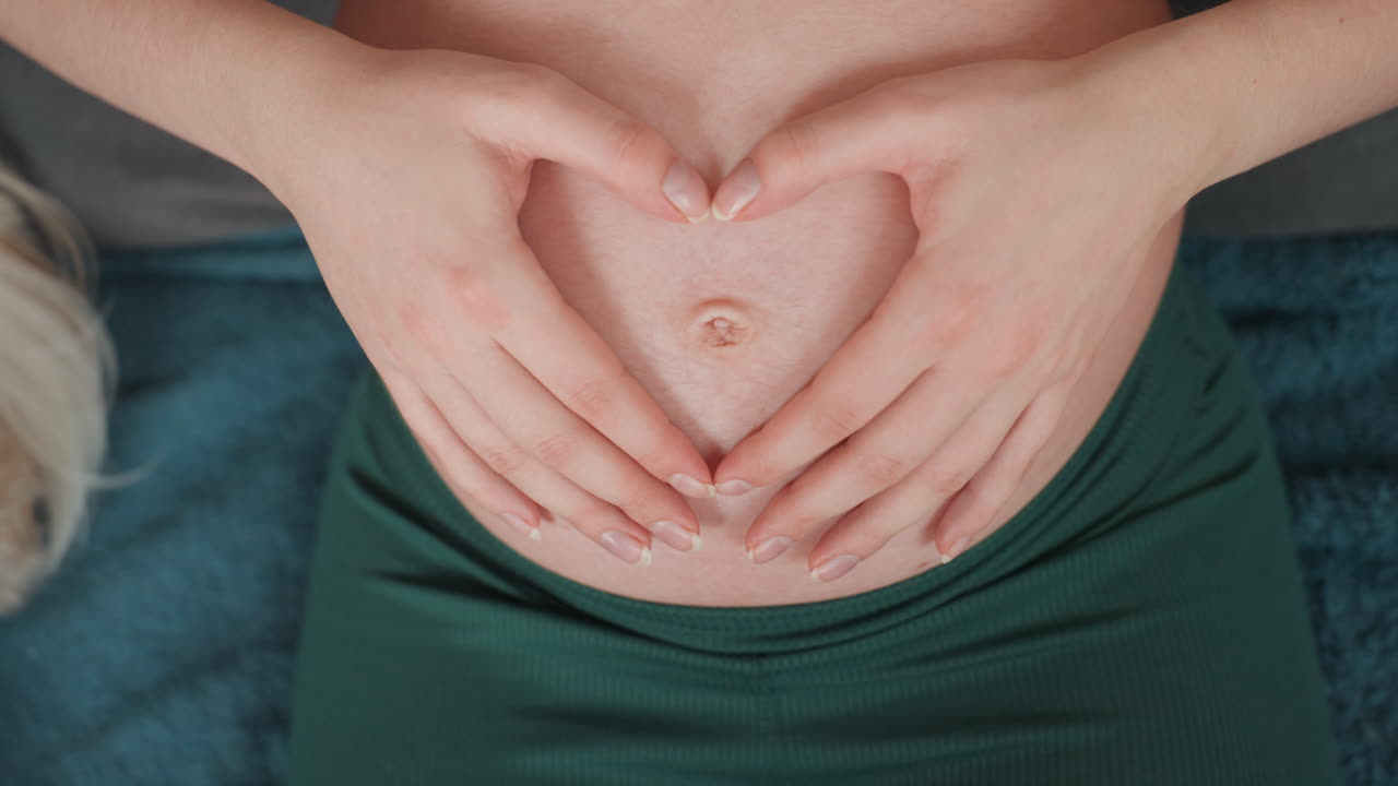 Maternity heart gesture woman bump, mothertobe forming heart with hands over belly in green dress, soft textiles and serene lighting, nursery planning mood, tender connection and joyful anticipation