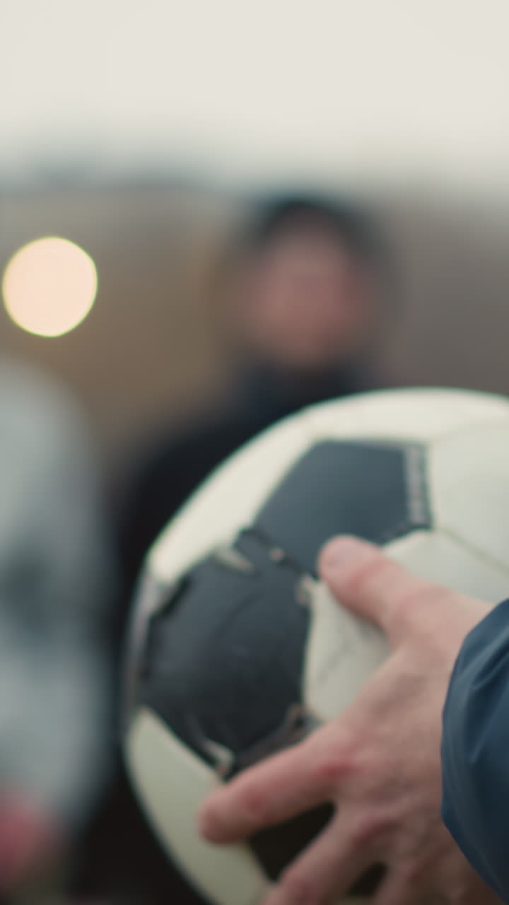 Close-up of a coach holding and turning a soccer ball in his hands, with a blurred view of two people standing in the background