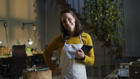 caucasian female jeweller in workshop wearing apron, holding tablet, smiling at camera