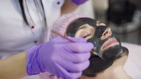 a young woman is lying on a couch with a black mask on her face, surrounded by a beautician which applying it in a beauty salon. close-up.