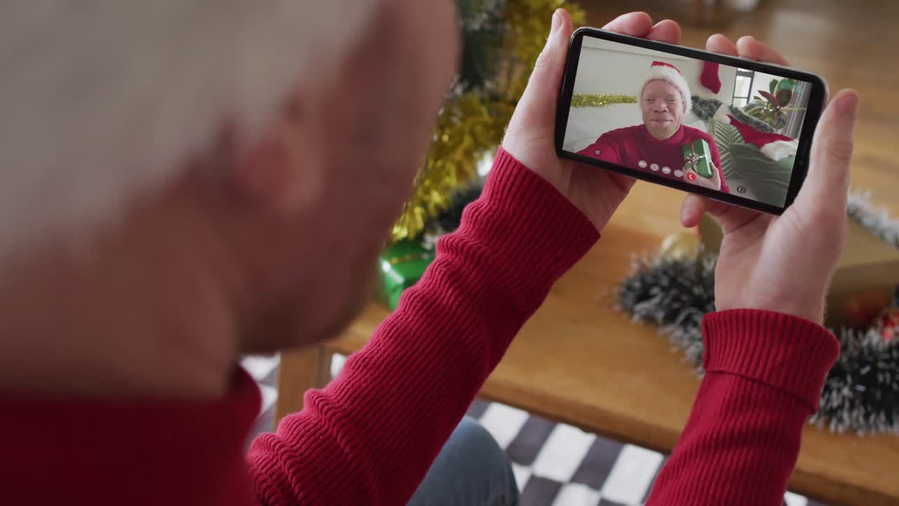 hombre caucásico con sombrero de santa usando un teléfono inteligente para una videollamada de navidad, con un hombre en la pantalla