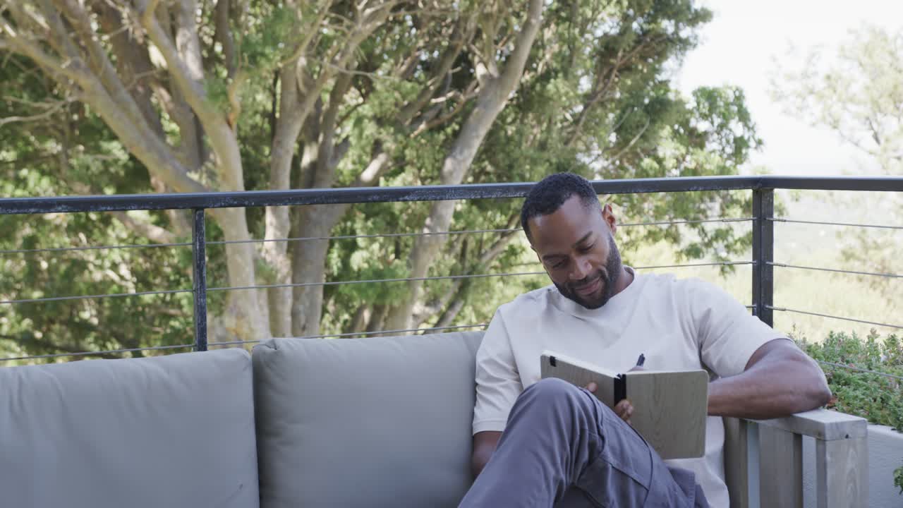African American man sitting on right balcony sofa using covered tablet with stylus, copy space
