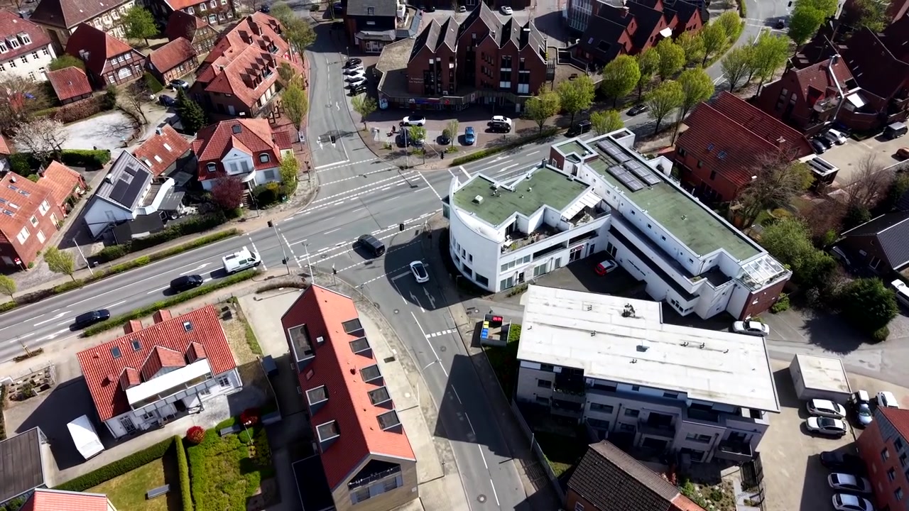 Luftaufnahme aus der Vogelperspektive eines deutschen Wohnviertels mit modernen Häusern, roten Dächern und einem weißen Tesla Elektroauto, das an einem sonnigen Frühlingstag in Europa durch die Straßen fährt.