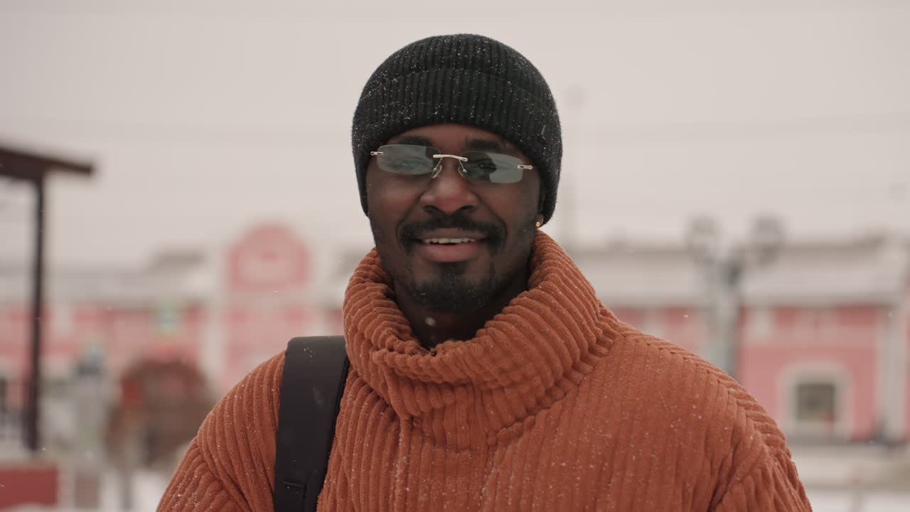 Uomo nero che sorride durante una passeggiata sulla neve, indossa un maglione di lana arancione e un berretto, occhiali e guanti, uno zaino in spalla, mentre parla al telefono in una piazza cittadina dai colori pastello con una leggera nevicata, un ritratto spontaneo.