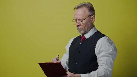 portrait of serious grey-haired man with a folder. senior man in glasses in suit writing on a folder. old man expresses different facial emotions on yellow background.
