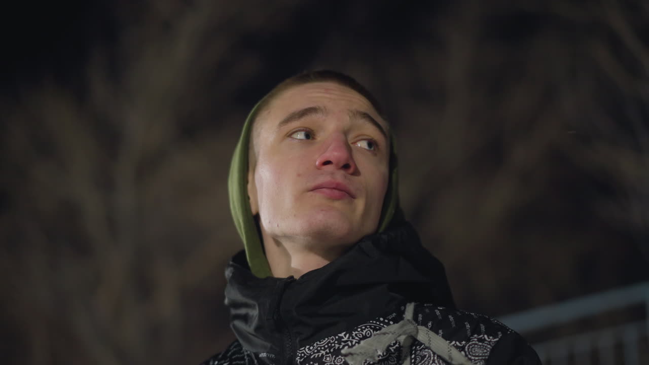 Close up of man in hooded jacket looking around thoughtfully on cold night, visible breath escaping into dark sky, bare trees forming soft background under dim winter lighting