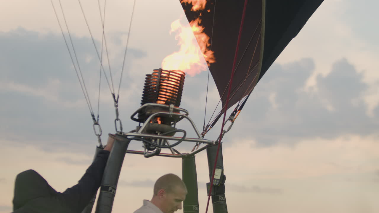Rear view of man wearing gloves adjusting metal frame and burner system during hot air balloon setup while another person in white cap assists nearby under soft morning sky with calm light