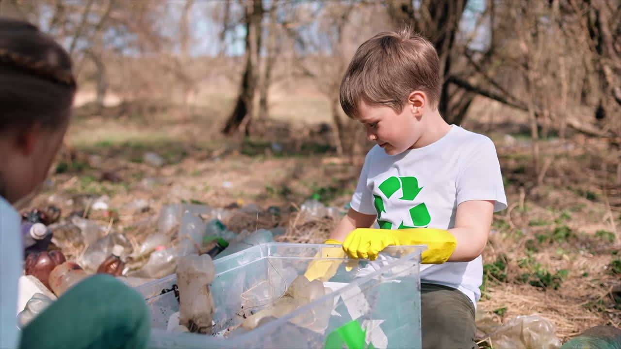 Boy and girl collecting plastic garbage in a container in a polluted clearing, recycling signs on the T-shirts. Slow motion