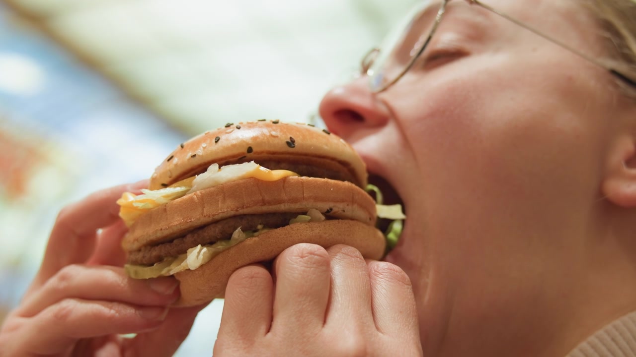 Closeup white woman taking big bite, food blogger persona with oversized sesame burger and fried egg, enthusiastic expression, hands gripping bun, bright mall interior, casual craving shot