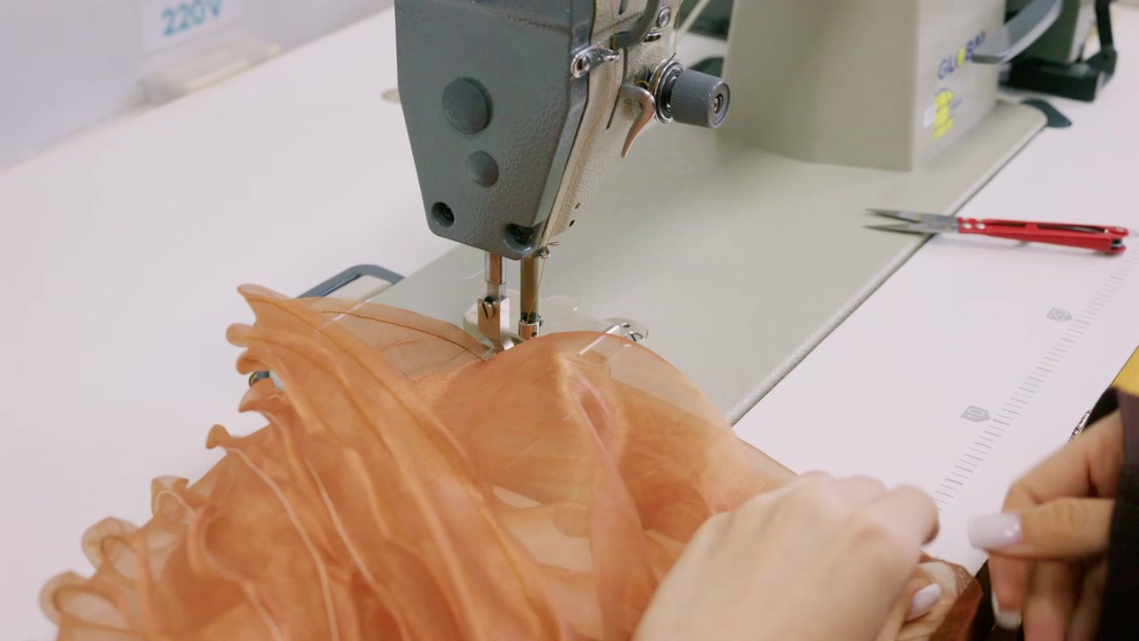 Uno stilista guida un tessuto di organza arancione traslucido sotto l'ago di una macchina da cucire industriale