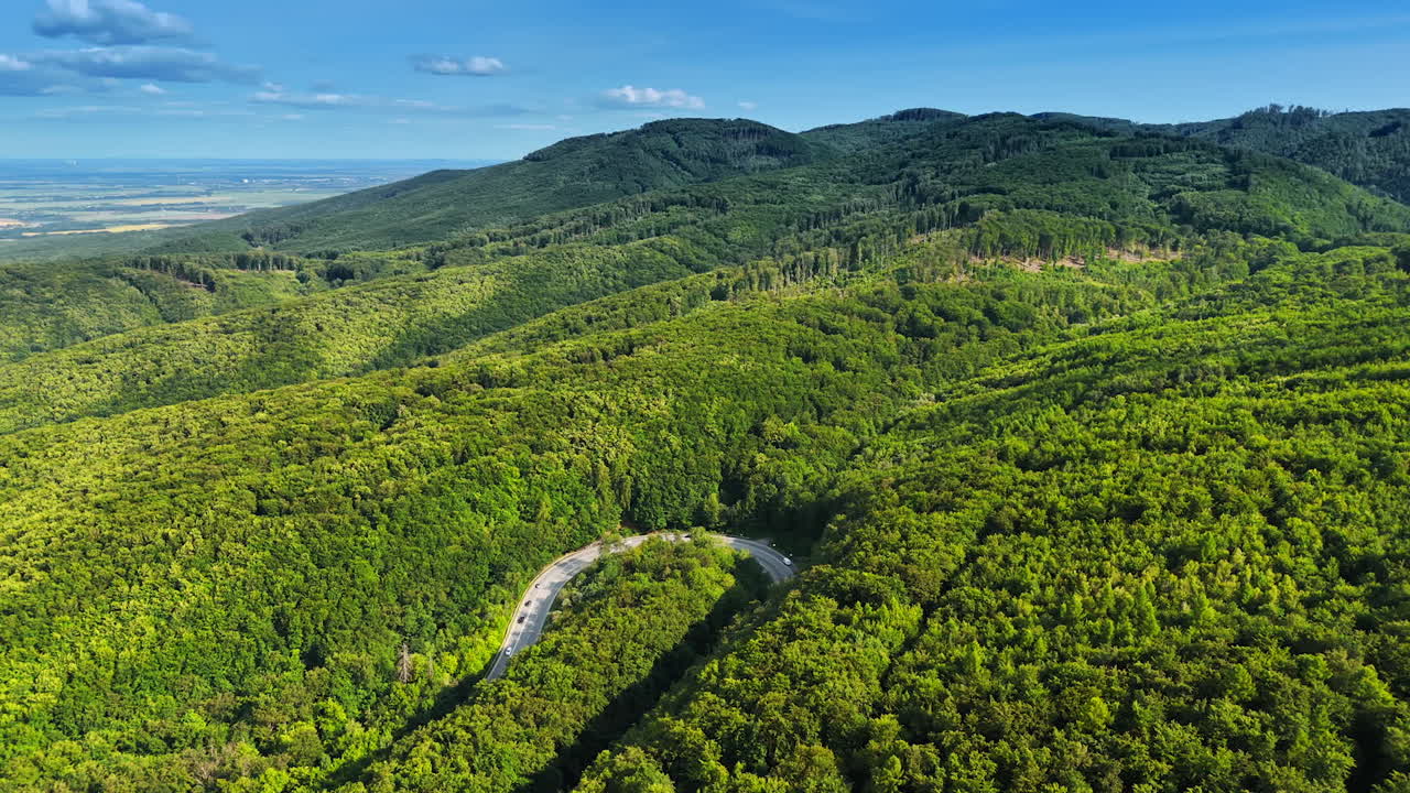 Loop of the highway crossing the lush green forests. Verdant mountain slopes at the backdrop of blue sky. Aerial view.