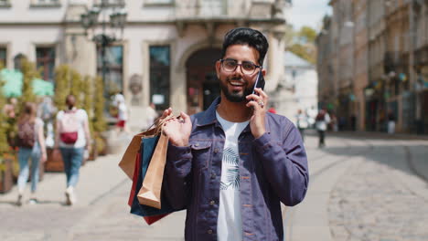 happy young indian man shopaholic consumer after shopping sale with full bags walking in city street