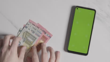 disparo de moneda por encima de la cabeza persona contando billetes y monedas de euro junto a un teléfono móvil con pantalla verde