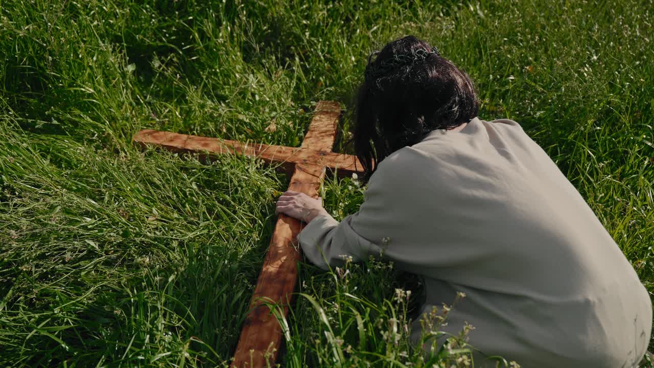 Pessoa ajoelhada na grama segurando uma cruz de madeira simbolizando o sofrimento de Jesus e a fé cristã durante a reflexão da Paixão de Cristo.
