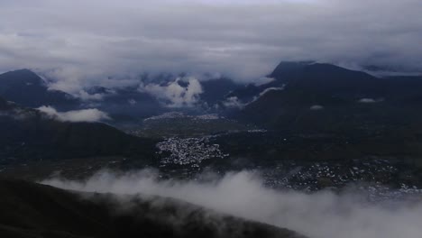 dark, gloomy aerial shot as flying over a ridge fog whirling around as clouds covering mountain top around a mall city in the valley