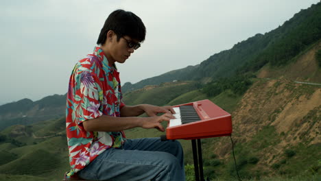 Músico-Asiático-Masculino-Toca-El-Piano-En-El-Paisaje-Montañoso-Natural-Con-Jeans-Vintage,-Camisa-Hawaiana-Y-Gafas-De-Sol