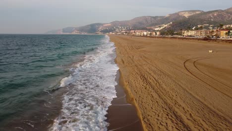 Playa-De-Castelldefels-En-Barcelona-Con-Olas-Bañadas-En-Tierra,-Playa-De-Arena-Vacía,-Telón-De-Fondo-De-La-Ciudad-Costera,-Vista-Aérea