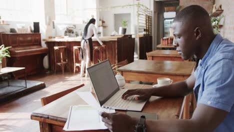 serious african american man using laptop and doing paperwork at coffee shop, slow motion